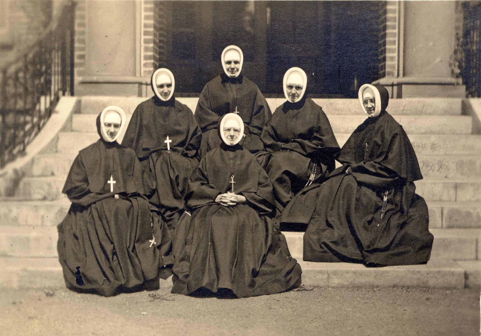 six Sisters of Providence from 1916 are seated on concrete steps in full habit
