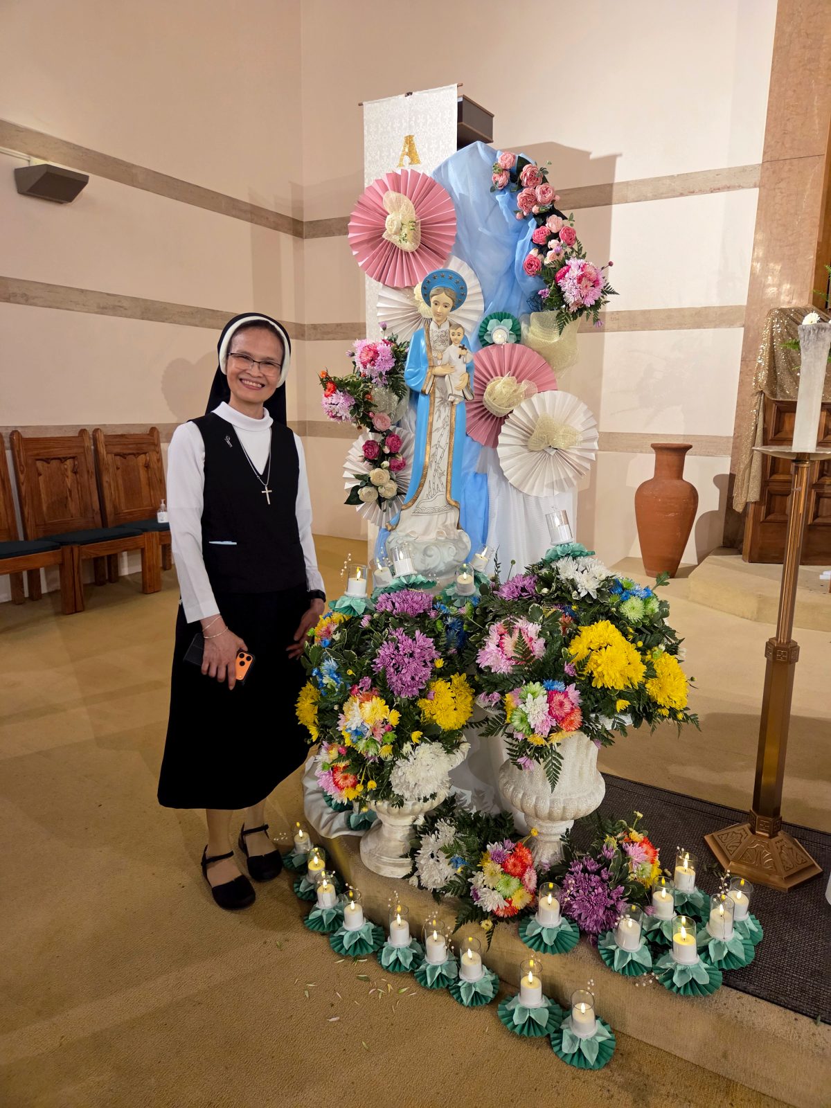 Una religiosa posa junto a una estatua de la Santísima Virgen María, adornada con flores.
